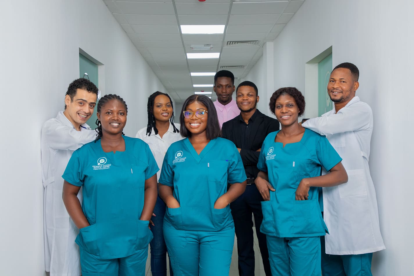 About A diverse medical team at the German Ozone Medical Centre posing in a bright hallway. The team consists of professionals in teal medical scrubs, white lab coats, and business attire, all smiling confidently.